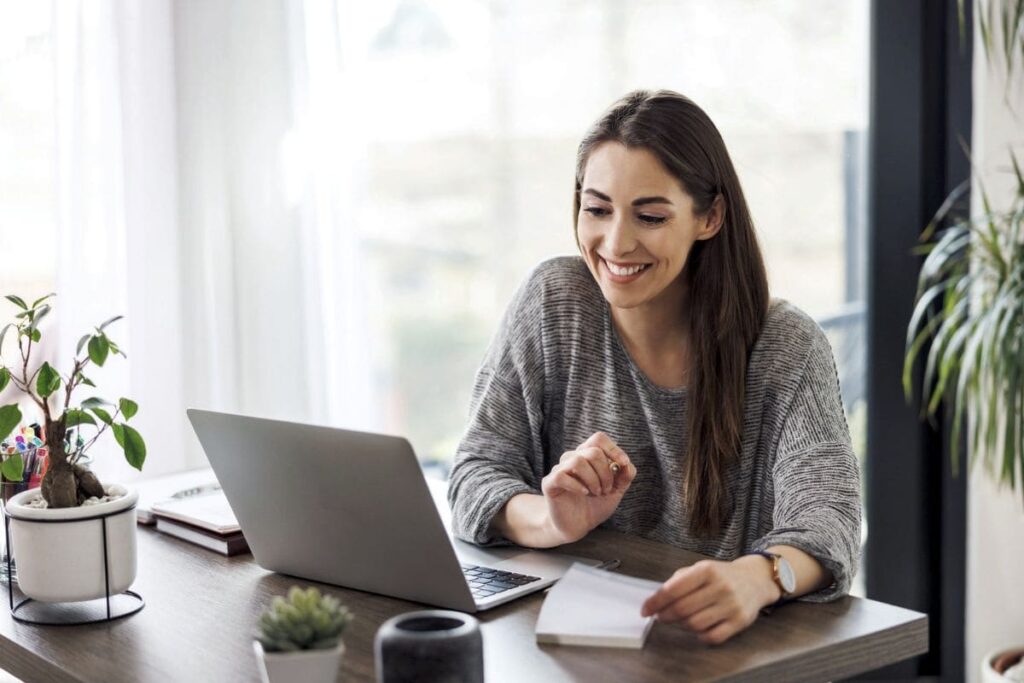 A financial planner, sitting at her home office desk, using a computer and calculator while writing in a notebook.