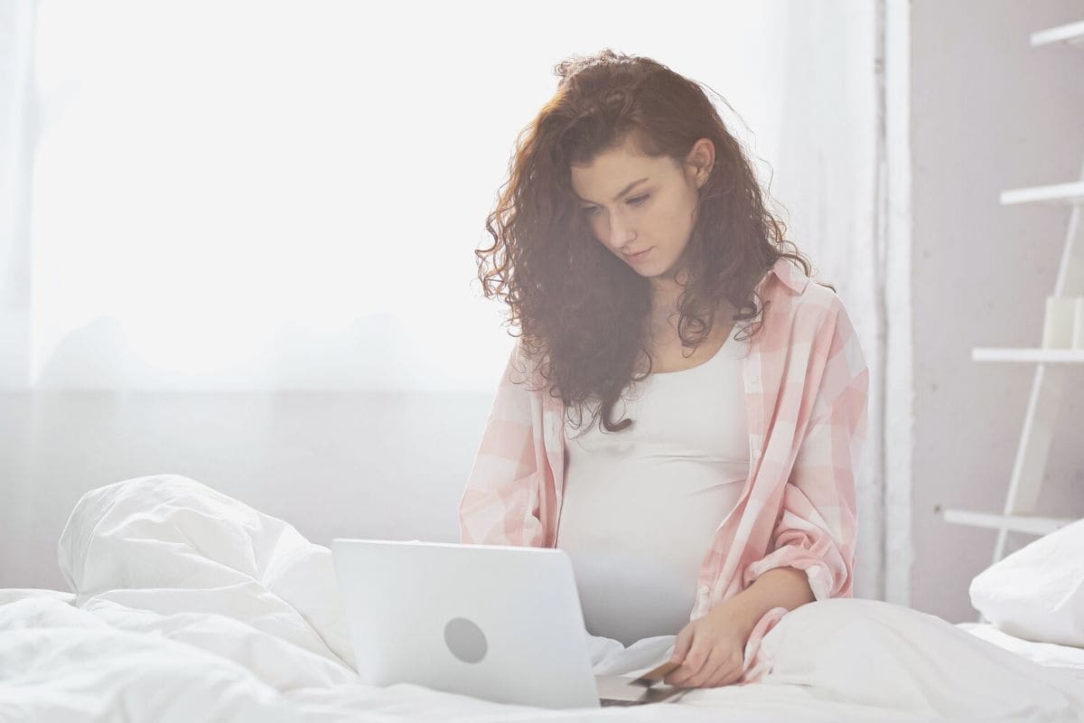 A pregnant woman sitting in bed and using a laptop to work at home.