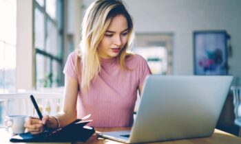 A Content Moderator working remotely from home, using a laptop and writing notes in a notebook for blog post content moderator jobs