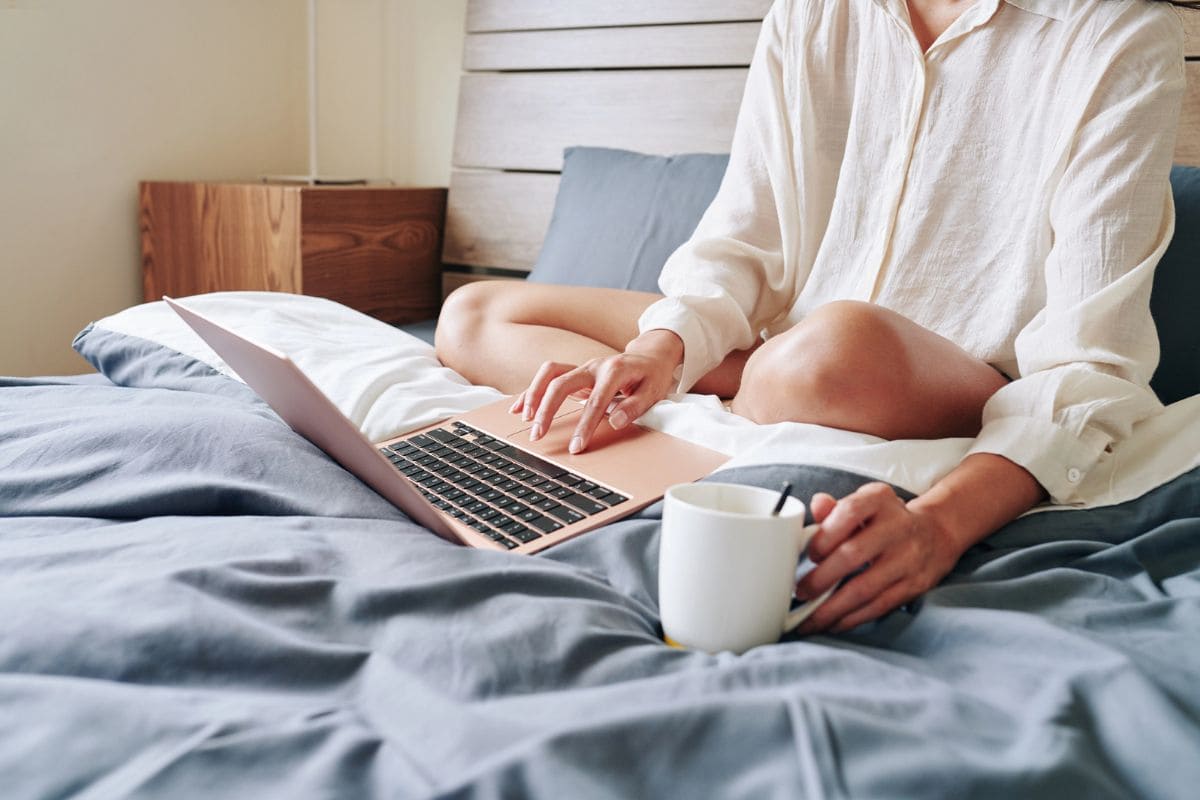 A woman working from home, using her laptop in bed while wearing pajamas for blog post dream work from home jobs.