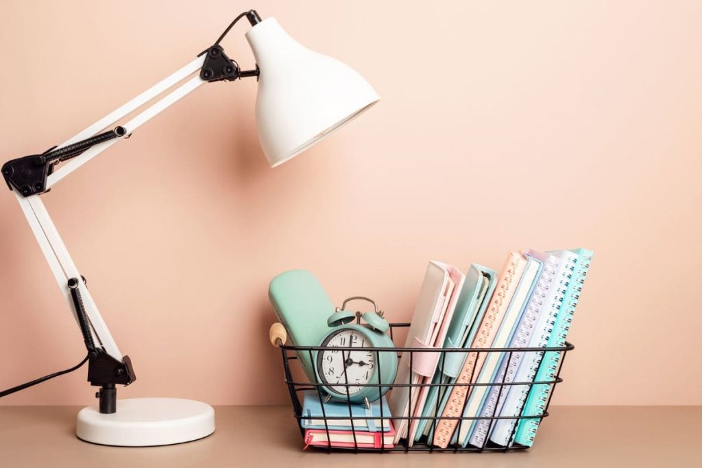 A teacher's home office desk with a lamp and basket of notebooks and school supplies.