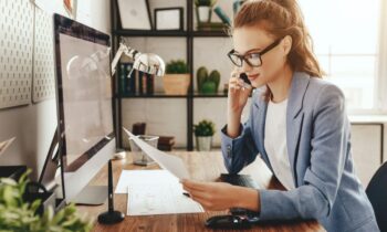 A woman working remotely from her home office, using a desktop computer and talking on a cell phone for blog post high paying entry-level jobs