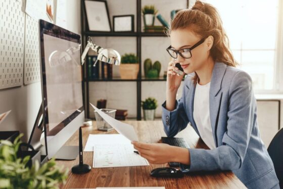 A woman working remotely from her home office, using a desktop computer and talking on a cell phone for blog post high paying entry-level jobs