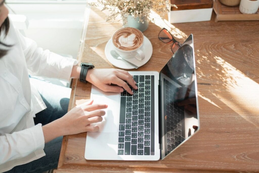 A woman working remotely from a cafe, using a laptop and drinking a cup of coffee.