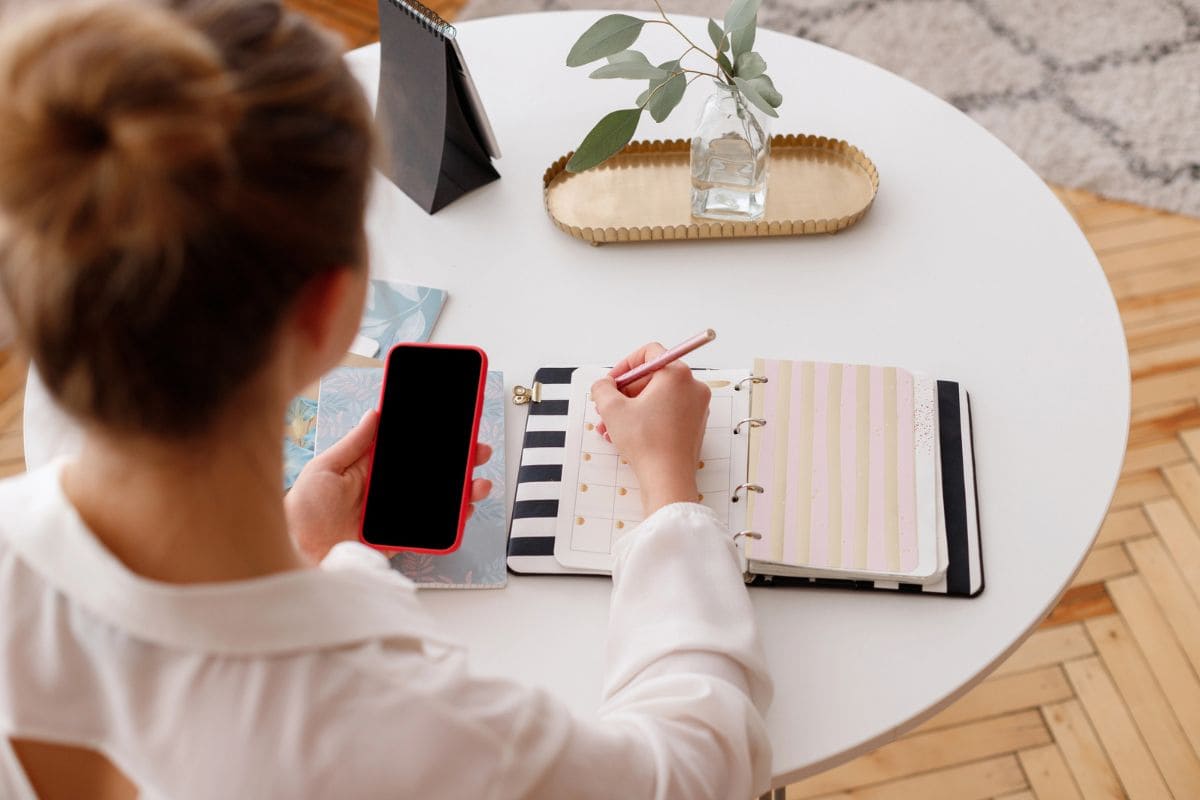 Overhead view of a woman using a smartphone and writing in a notebook at a desk for blog post top-rated gig apps