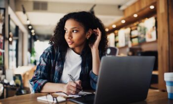 A woman sitting in a cafe, looking out the window, while holding a pen and writing in a notebook for blog post get paid to write poetry online