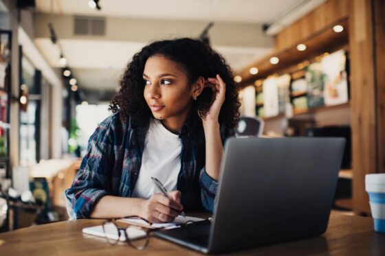 A woman sitting in a cafe, looking out the window, while holding a pen and writing in a notebook for blog post get paid to write poetry online