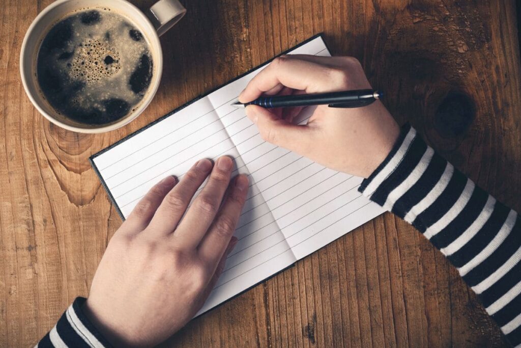 Una vista aérea de un escritor sentado en el escritorio de una oficina en casa, escribiendo en un cuaderno y tomando café.