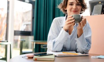 A woman working from her home office, sitting at a desk, writing in a notebook, and smiling while drinking coffee.