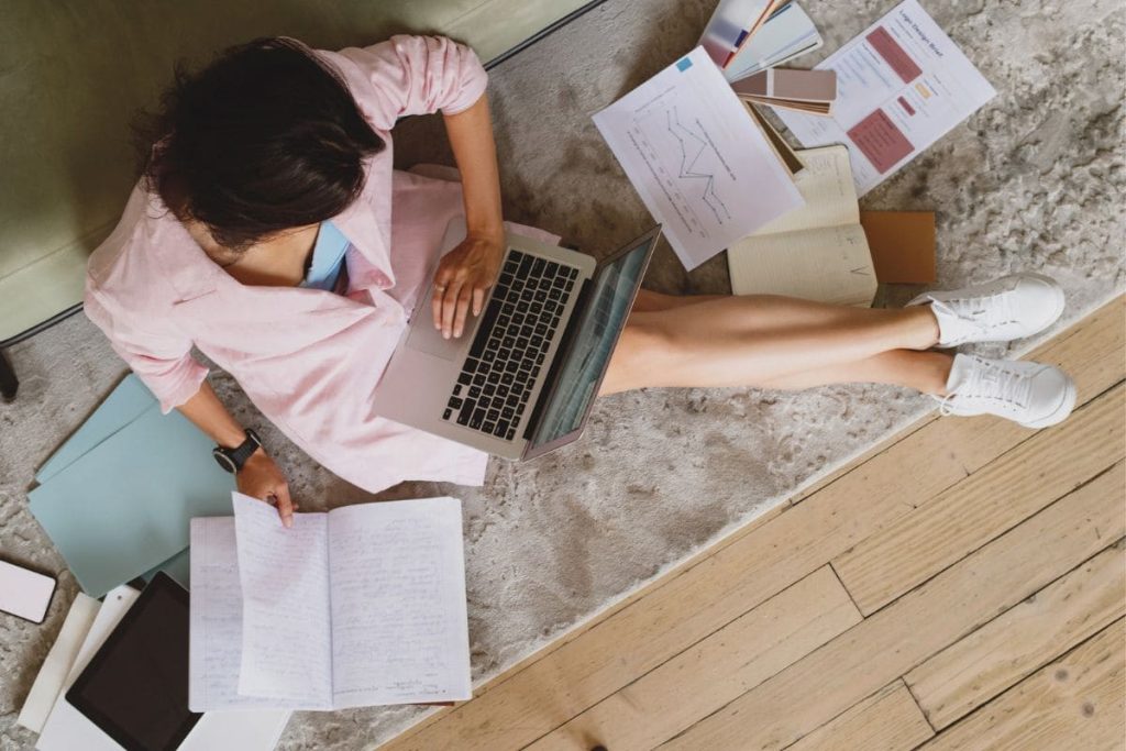 A woman working from home while sitting on the floor, using a laptop, surrounded by papers.