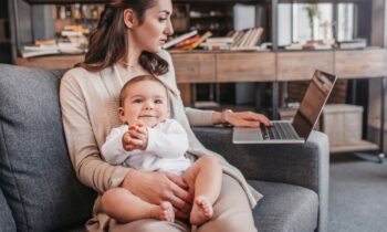 A mom holding her baby, while sitting on the couch and using a laptop for blog post family-friendly jobs for parents