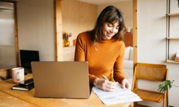 A woman working remotely from her home office desk, using a laptop and writing in a notebook.