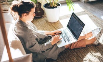 A woman sitting on the floor in her living room, working remotely on a laptop for blog post remote jobs with little human interaction.