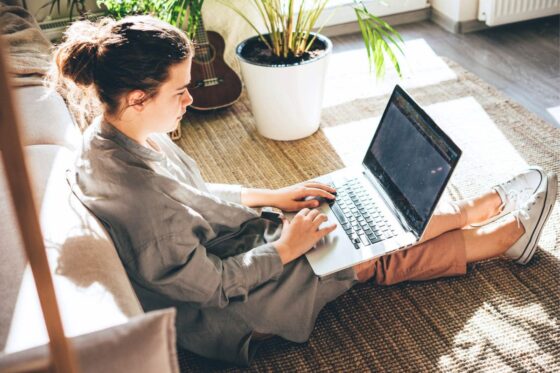 A woman sitting on the floor in her living room, working remotely on a laptop for blog post remote jobs with little human interaction.