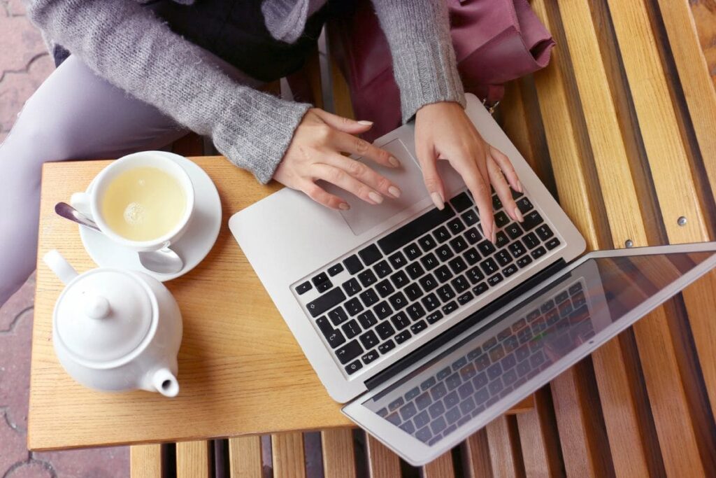 Close-up image of a woman's hands working remotely, drinking tea and typing on a laptop.
