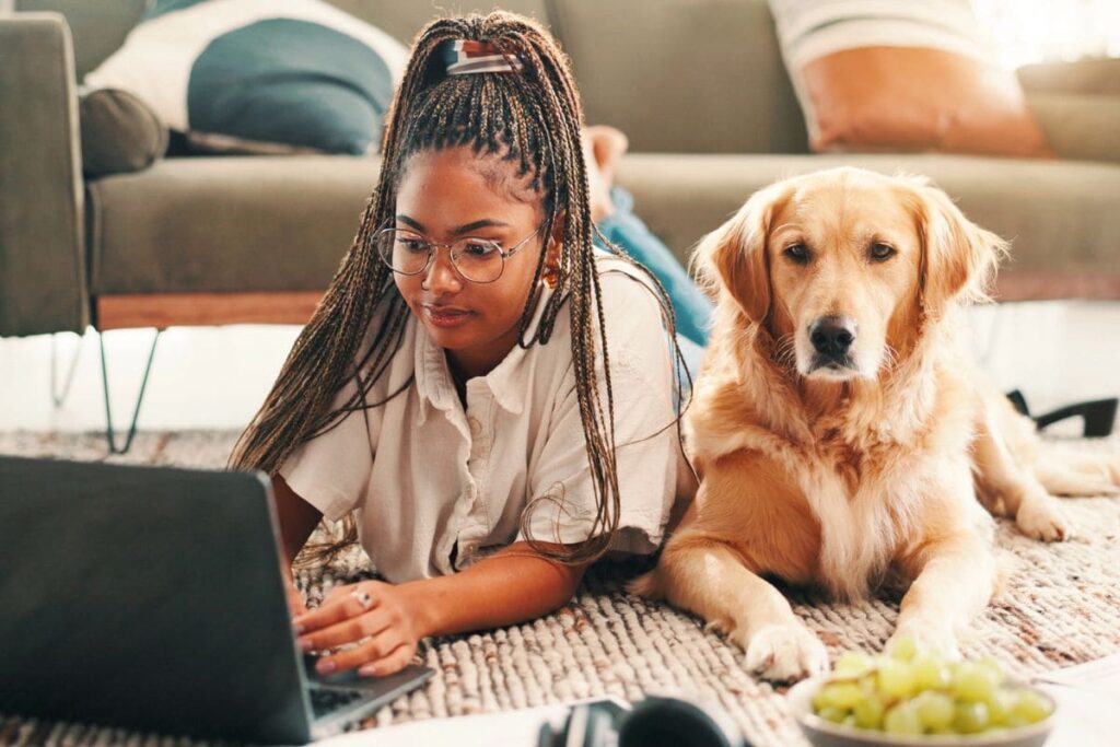 A woman working from home, lying on her living room floor, using a laptop, with a golden retriever sitting next to her.