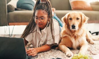 A woman working from home, lying on her living room floor, using a laptop, with a golden retriever sitting next to her.