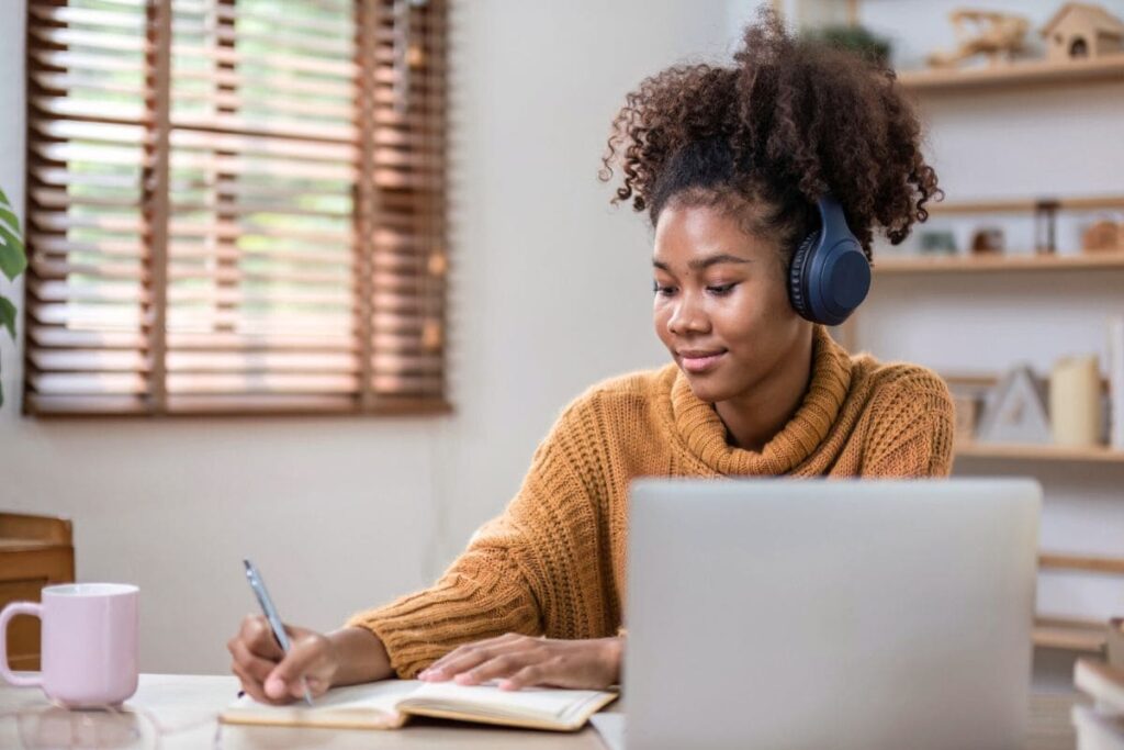 13 Easy Side Hustles for College Students to Earn Extra Cash 3 College student working from home, using a laptop, wearing headphones, and writing in a notebook.