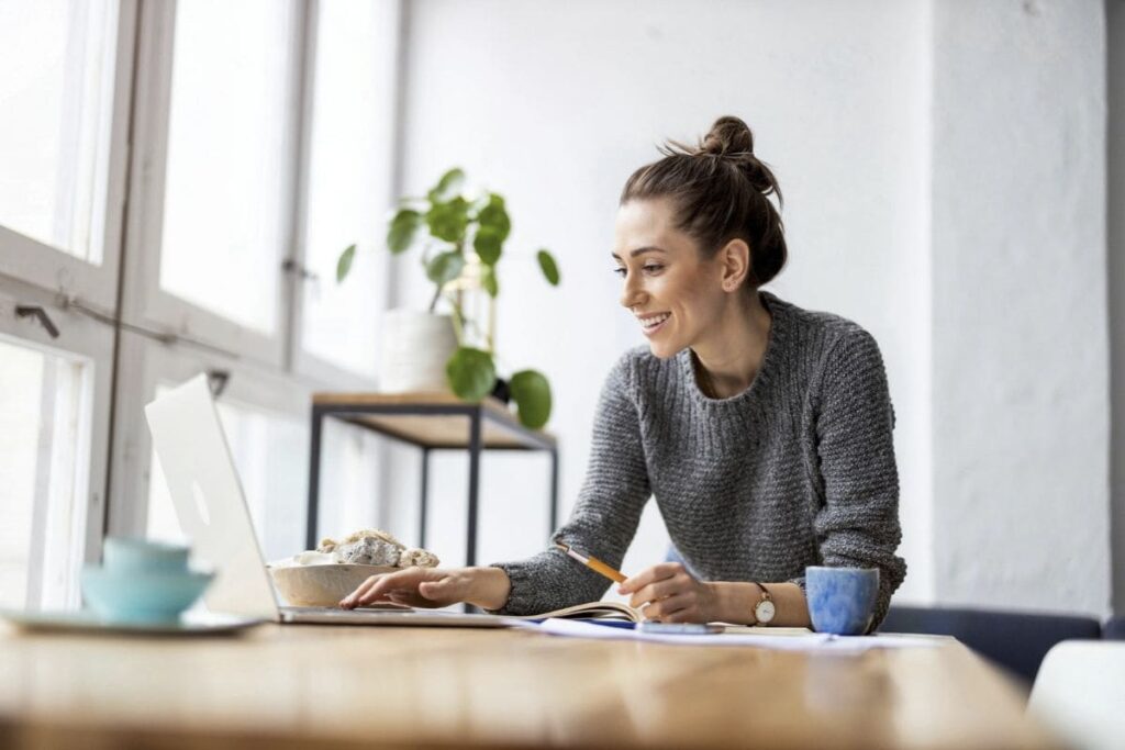 A remote worker smiling, while sitting at her desk, using a laptop.