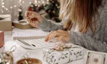 A woman creating a holiday budget, using a calculator and writing in a notebook while surrounded by holiday decor for blog post save money this holiday season