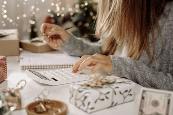 A woman creating a holiday budget, using a calculator and writing in a notebook while surrounded by holiday decor for blog post save money this holiday season
