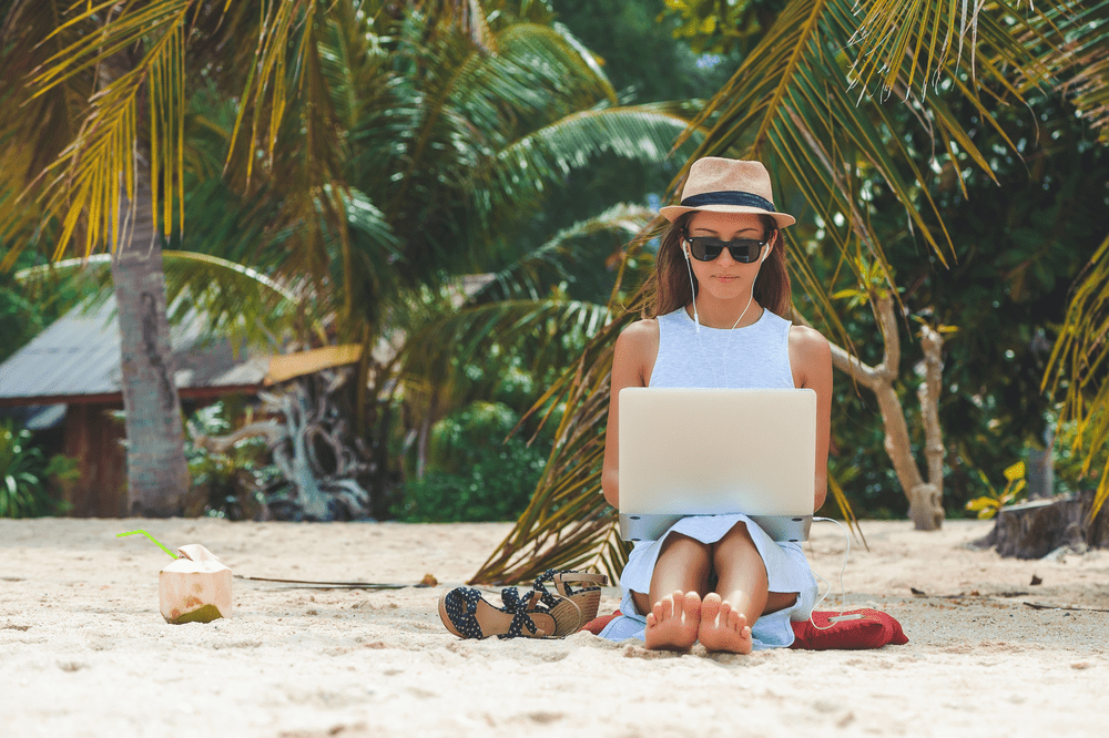 woman working on the beach with her laptop wearing a hat and sunglasses