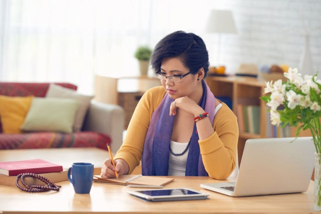 A woman working from home at a desk and writing in a notebook.