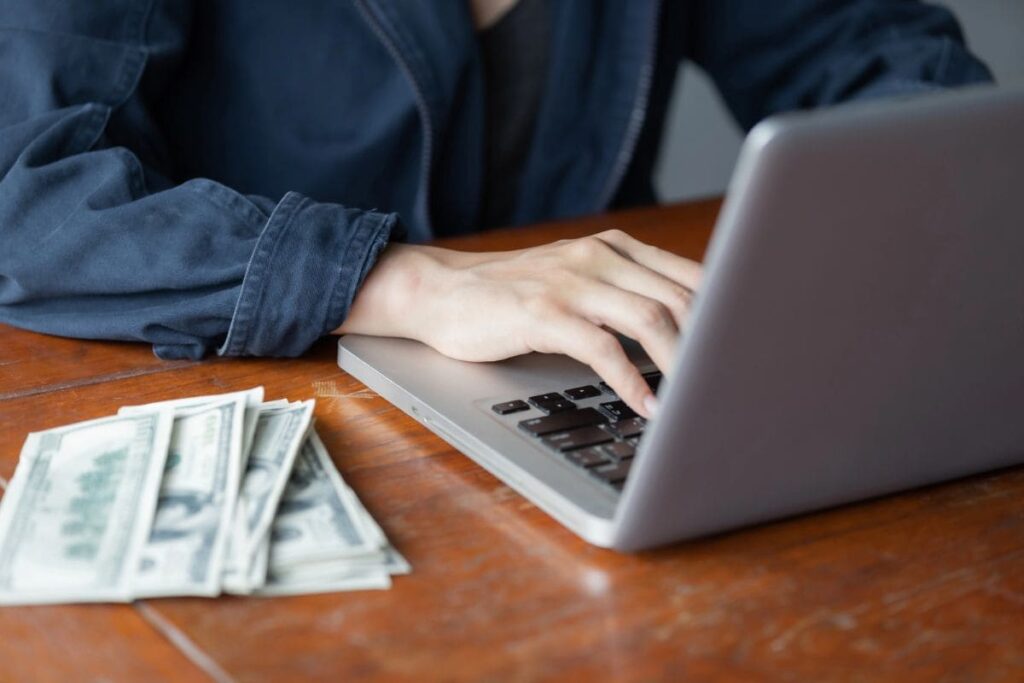 A woman working on a laptop at her home office desk, with cash sitting on the desk beside her.