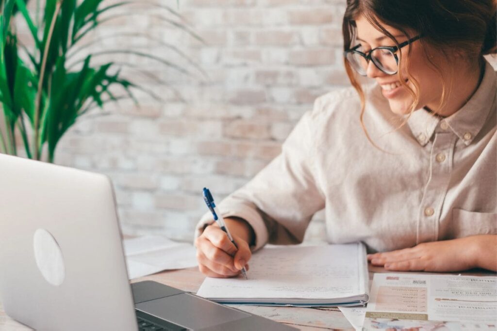 A woman working remotely from her home office, using a laptop and writing in a notebook.