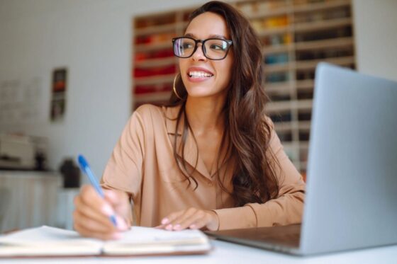 A woman working remotely from her home office, using a laptop and writing in a notebook.