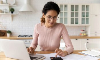 A woman working remotely from her kitchen table, using a laptop and a calculator for blog post work from home jobs with paid training