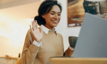 A woman using a laptop in her home to take a video interview call, smiling and waving at the camera for video job interview tips
