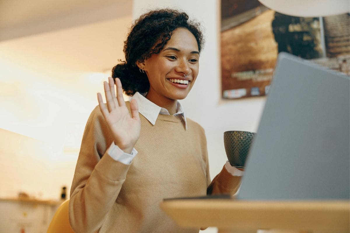 A woman using a laptop in her home to take a video interview call, smiling and waving at the camera for video job interview tips