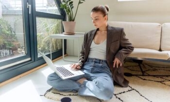 A woman sitting on the floor in her living room and working from home on a laptop.