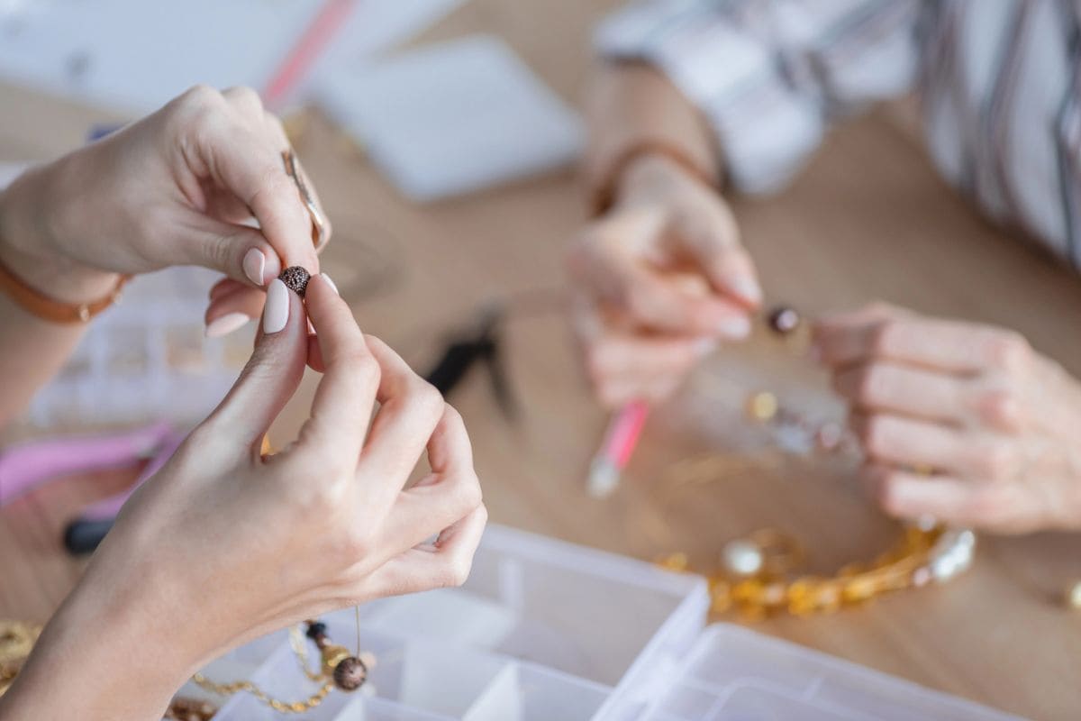 Closeup image of a woman's hands, holding beads and making jewelry at a large craft table.