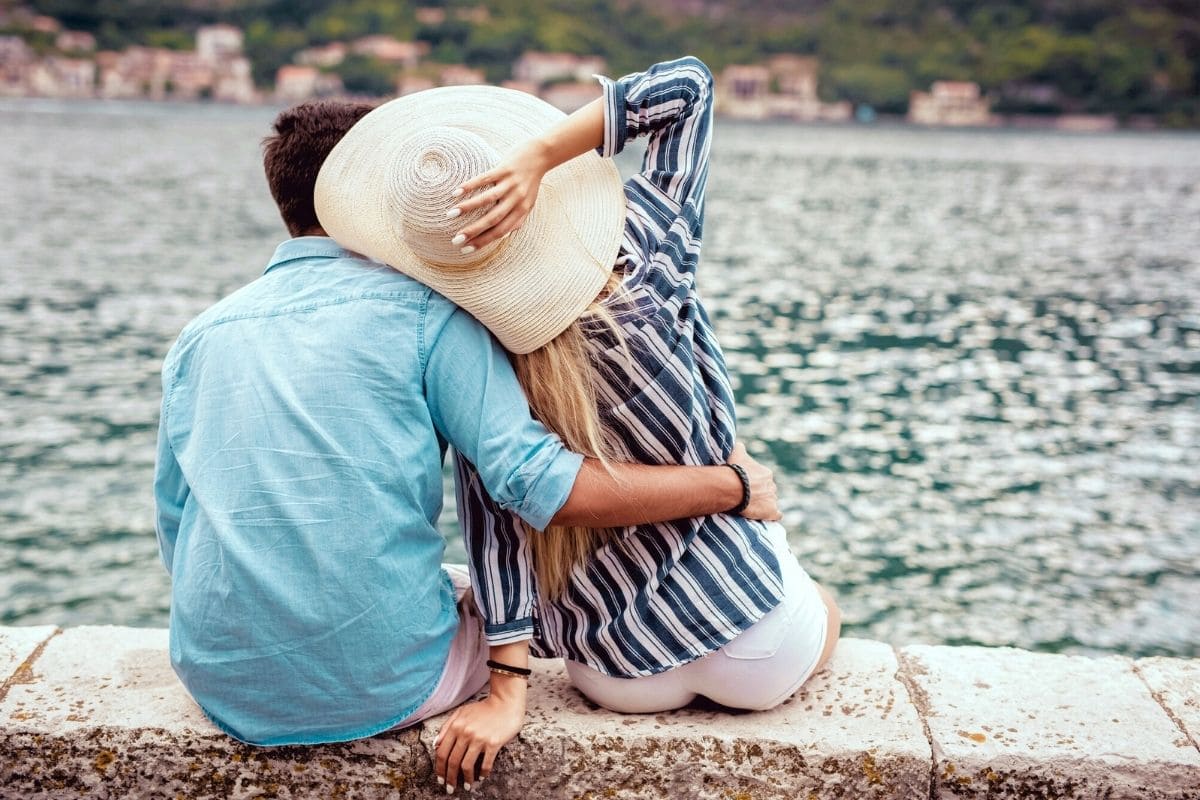 A couple sitting together on a rock wall, overlooking a lake with hills and buildings in the distance for blog post romantic business ideas