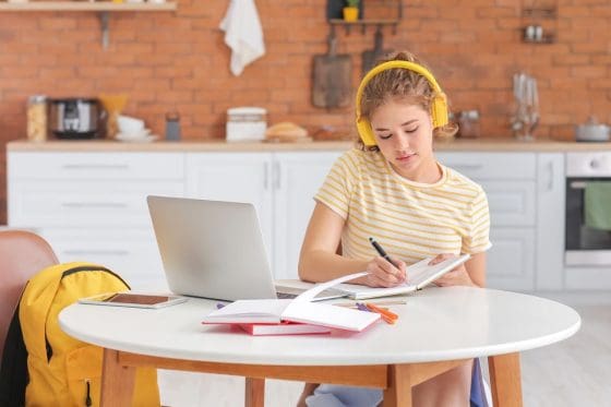 Young teen girl working at home on a laptop at the kitchen table, wearing headphones and writing in a notebook.