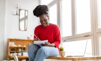 A woman working from home, sitting near a window, writing in a notebook.
