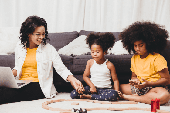 Mom sitting on floor with two daughters working on laptop while they play with their train set