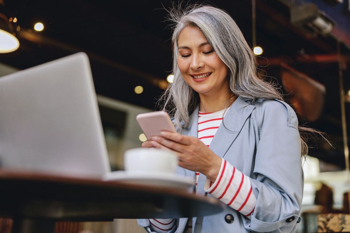 Woman sitting at a table in a cafe, using a smart phone and a laptop to work remotely for blog post work from home jobs that provide equipment
