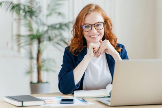 A woman business owner, smiling and sitting at her home office desk for blog post 50 service business ideas.