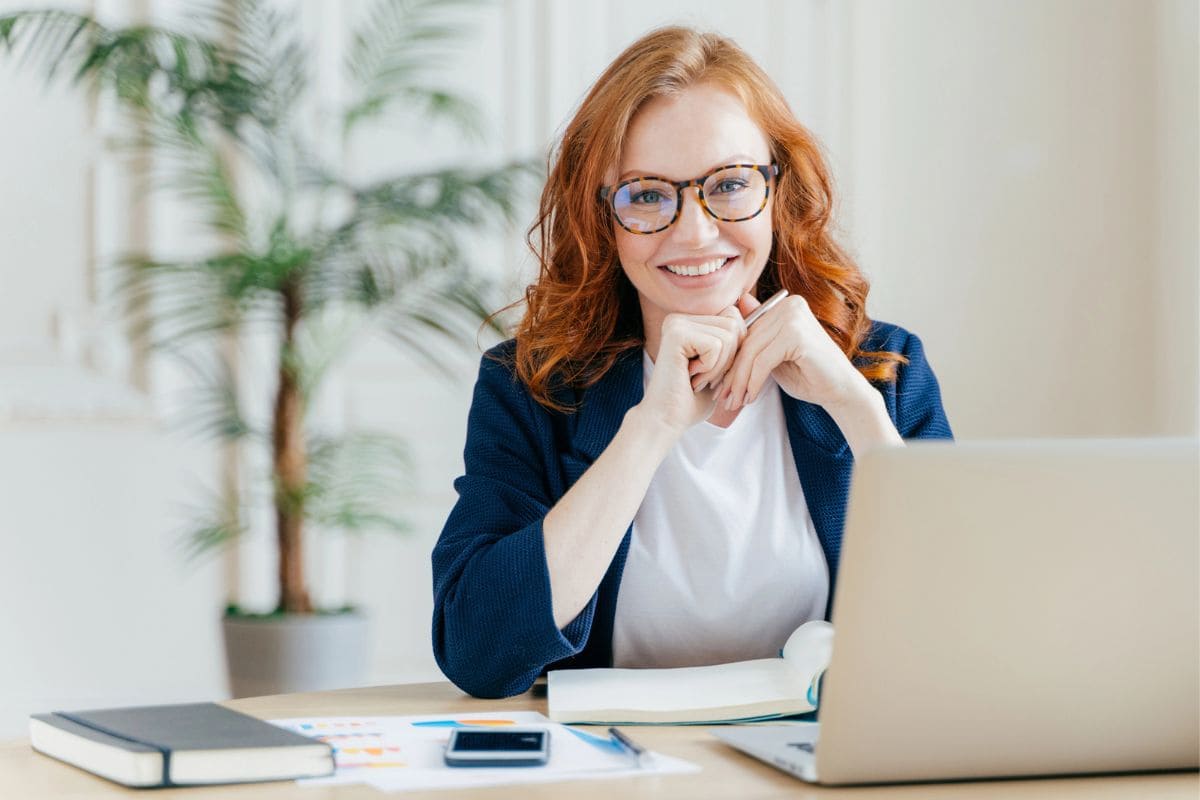 A woman business owner, smiling and sitting at her home office desk for blog post 50 service business ideas.