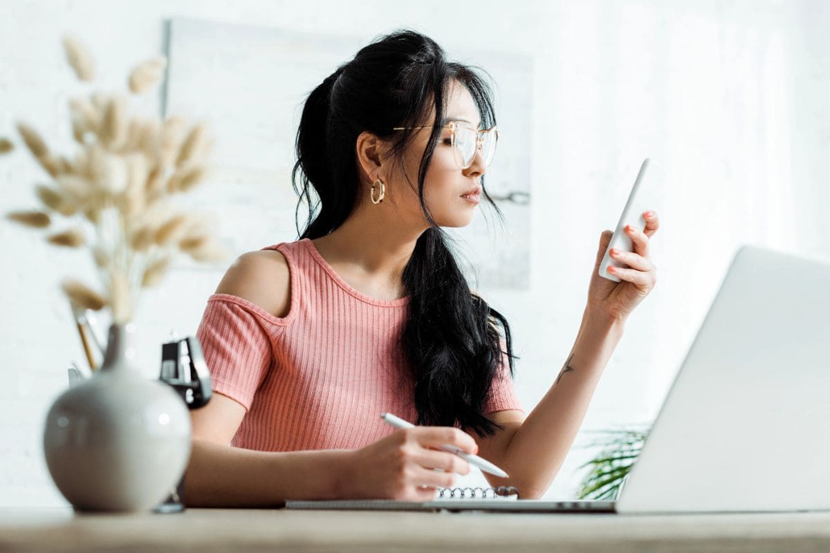 A woman working at a home office desk, holding a smart phone and writing in a notebook.