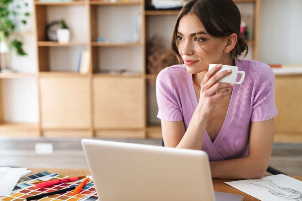 A woman working from home, sitting at a desk, using a laptop and drinking coffee.