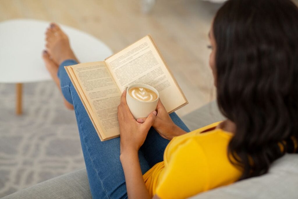 A woman sitting on the couch in her living room, reading a book, and holding a copy of coffee.