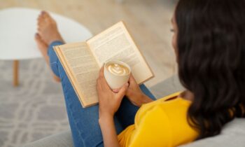 A woman sitting on the couch in her living room, reading a book, and holding a copy of coffee.