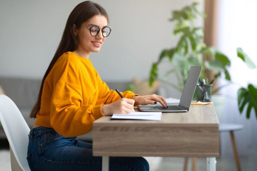 A woman with ADHD working at home using a laptop and writing in a notebook.