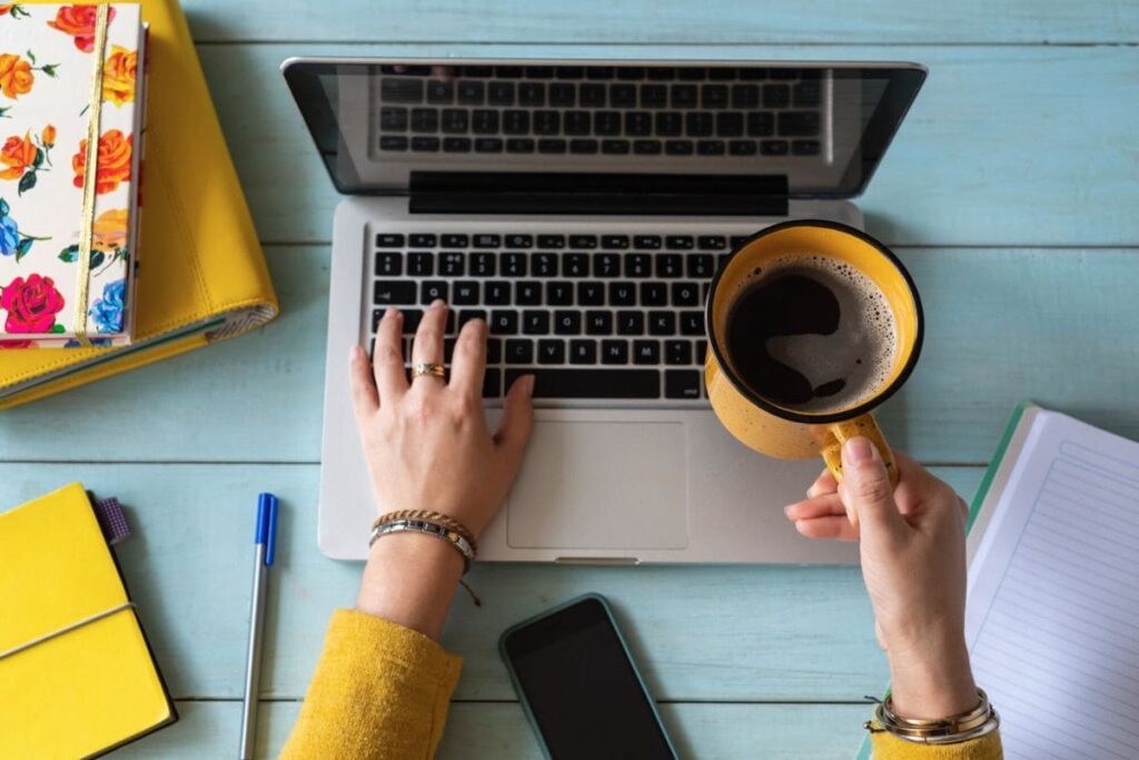 An overhead view of a woman working from a laptop at a table, drinking coffee.