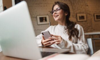 A woman working from a home office, sitting at a desk, holding a phone, and working on a laptop.