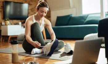 A woman getting ready to exercise at home, sitting in her living room on a yoga mat, putting on running shoes and using a laptop for blog post apps that pay you to exercise